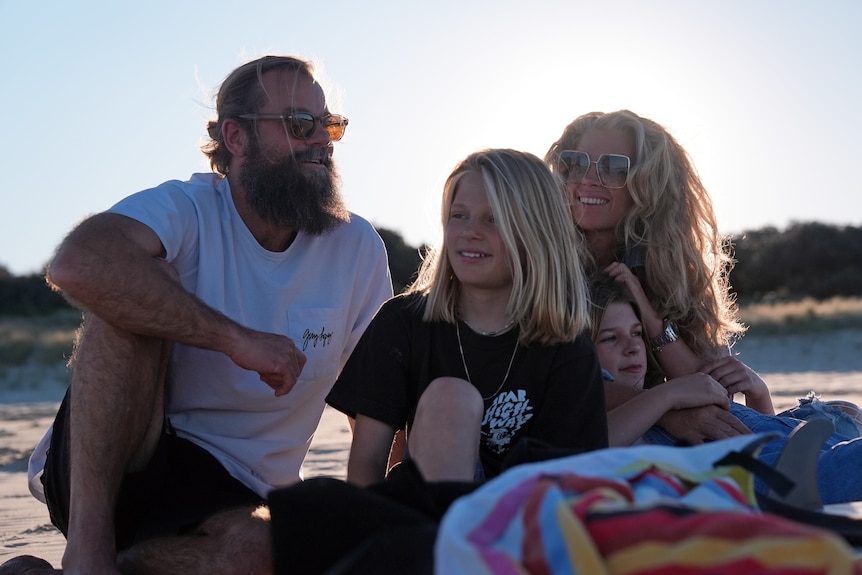 A man and woman smile sitting on the sand with their two kids