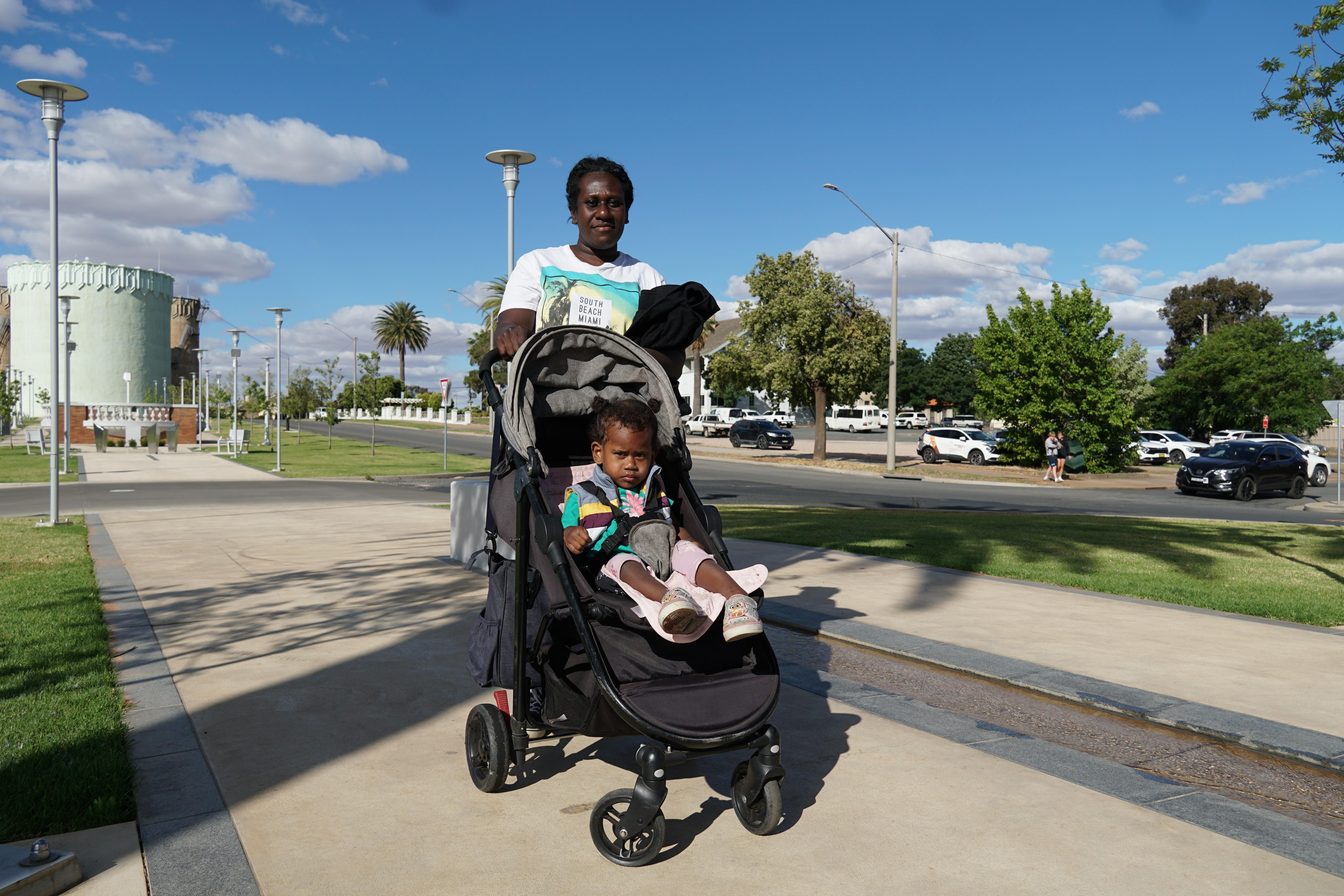A mother with a pram that carries her daughter, standing in a town centre with a water tower in the background.