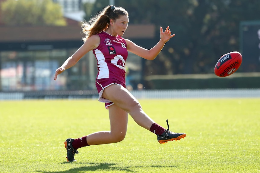 A young woman in maroon uniform kicking an AFL football.
