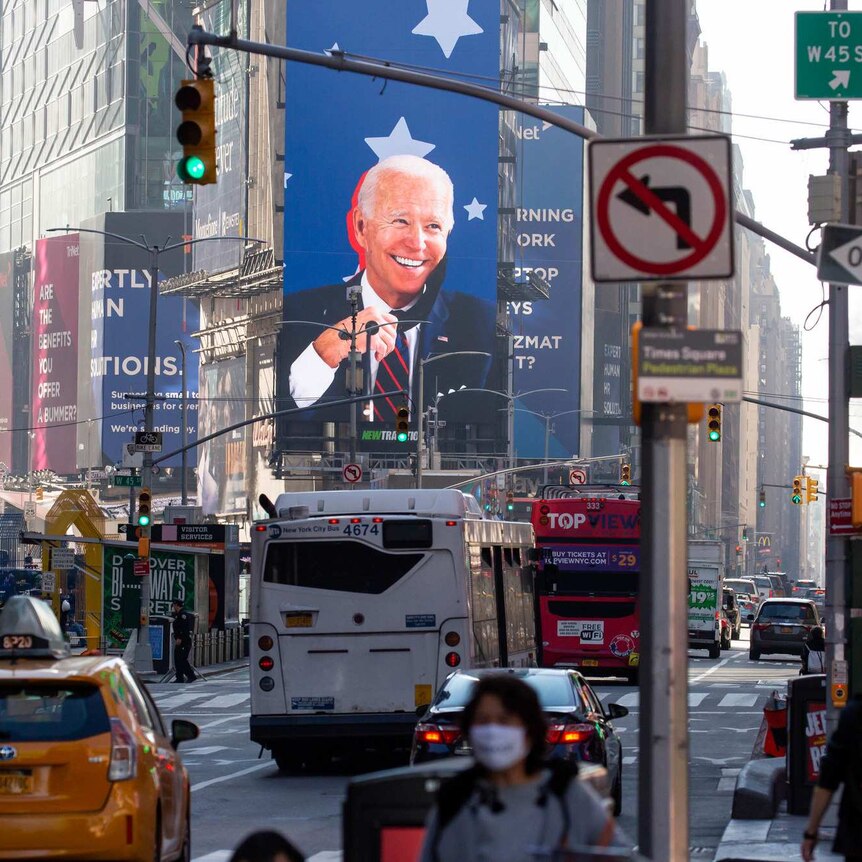 Joe Biden smiles from a large screen at Times Square in New York, as traffic and pedestrians pass by in the foreground.