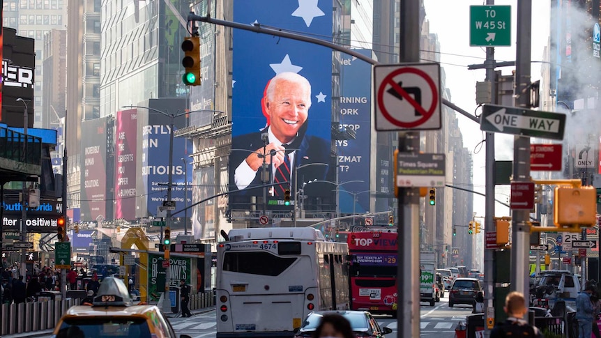 Joe Biden smiles from a large screen at Times Square in New York, as traffic and pedestrians pass by in the foreground.