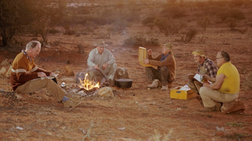 Five men sit around a campfire in a desert setting, painting.