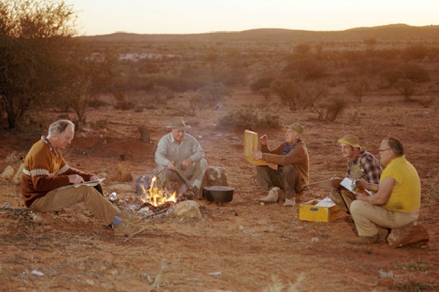 Five men sit around a campfire in a desert setting, painting.