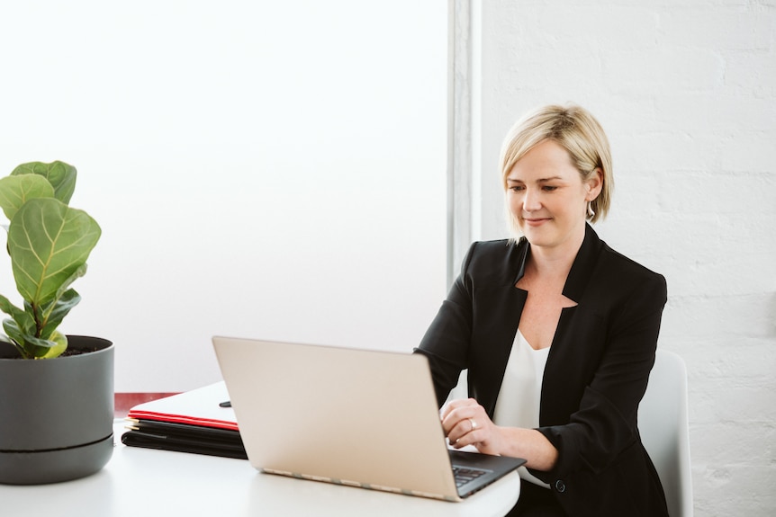 Caroline Grainger, who has a short blonde bob haircut and is wearing a smart blazer, types at a laptop.