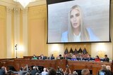 A woman with long blond hair addresses a room full of people via video link 