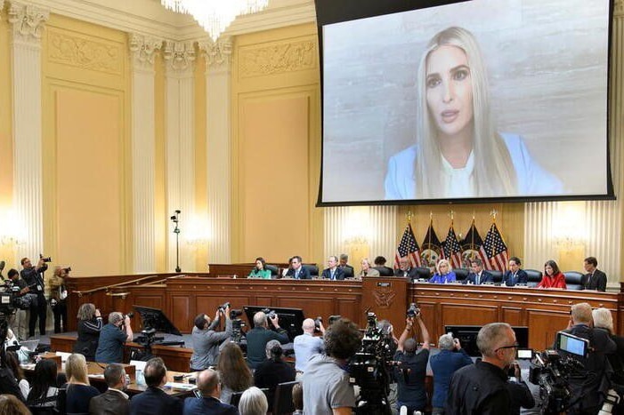 A woman with long blond hair addresses a room full of people via video link 