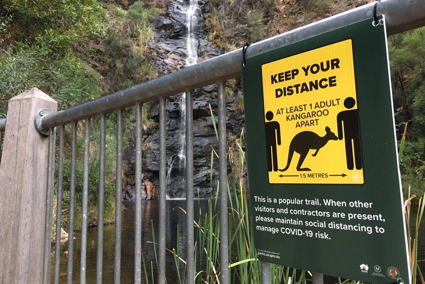 A fence with a sign saying to stay one kangaroo apart with a waterfall behind