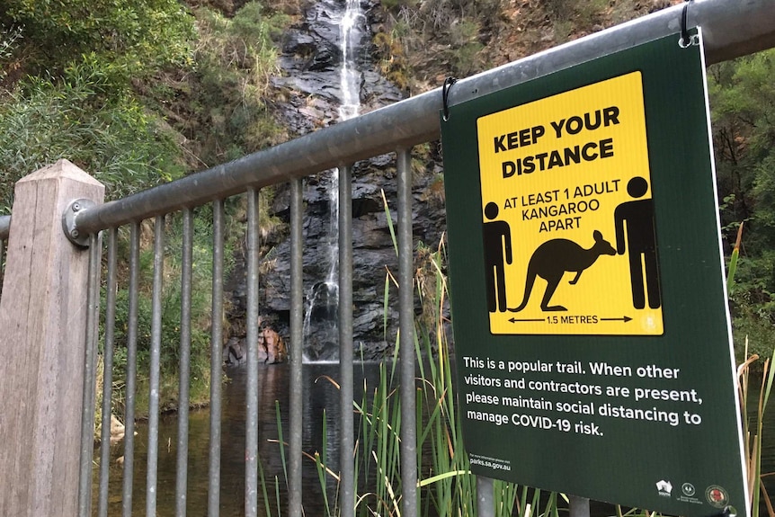A fence with a sign saying to stay one kangaroo apart with a waterfall behind
