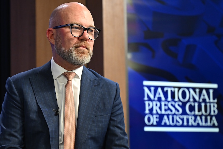 A bald, bespectacled man in a dark suit sits near a sign that reads "National Press Club of Australia".