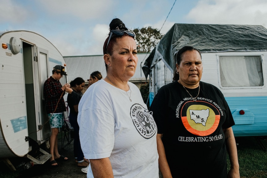 Two women stand in the foreground. Behind them are two caravans, a house and a few of their children.