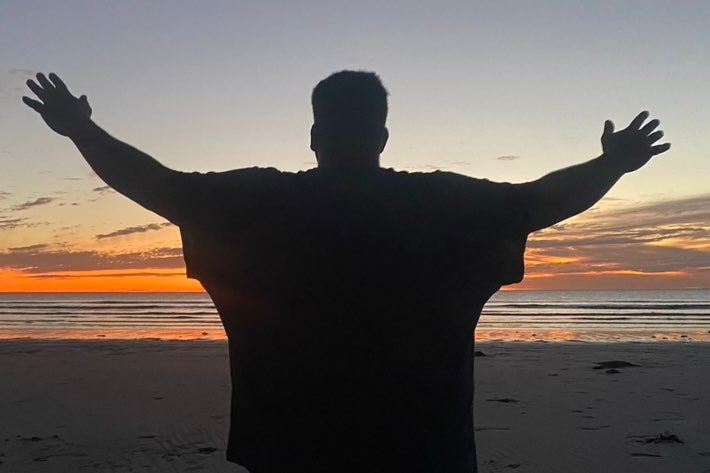 A man stands on a beach with his arms outstretched as he looks at the sunset. 