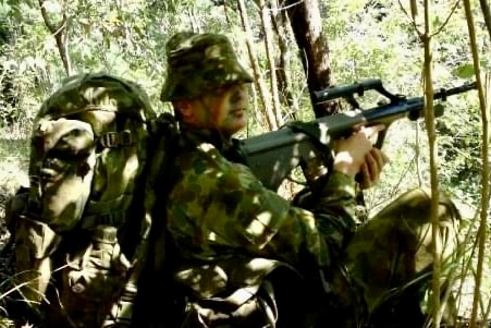 A man in combat fatigues with his face painted crouches in bushland, pointing a bullpup rifle.