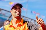 Man in a orange shirt and purple hat waving to a crowd outdoors.