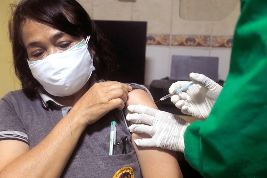 A woman wearing a mask lifts up her sleeve as another person injects a needle into her arm