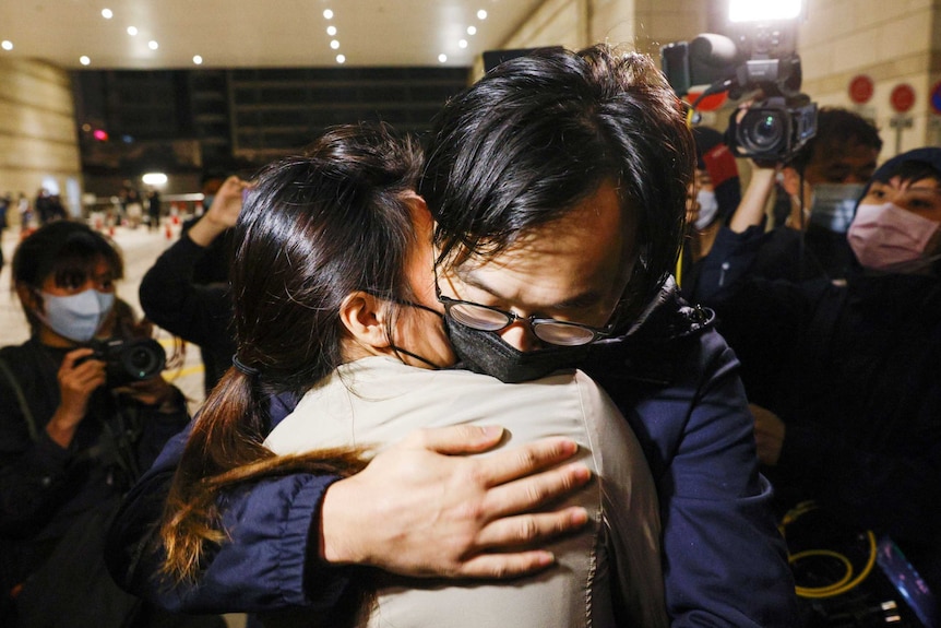 An Asian man in a face mask hugs a woman as photographers and journalists surround them.