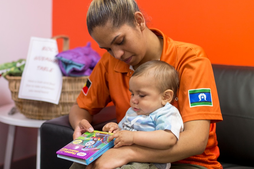 an image of an Indigenous mother and her bub sitting on a lounge with the baby holding a book