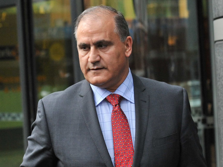 A man wearing a grey suit, red tie and blue shirt walking in front of a window.