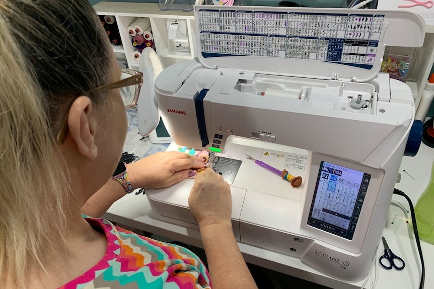 a woman sits at a table sewing a mask with a machine