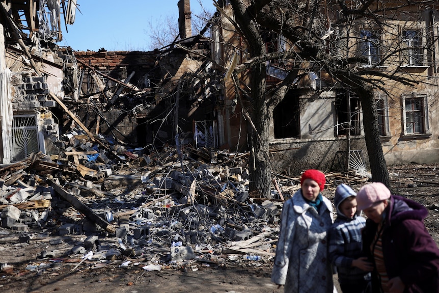 Two women and a child stand outside a house that appears to have been destroyed by an explosion. 