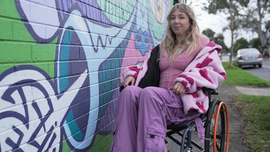 A young white woman with blonde hair sitting in a wheelchair, sat next to a mural on the street