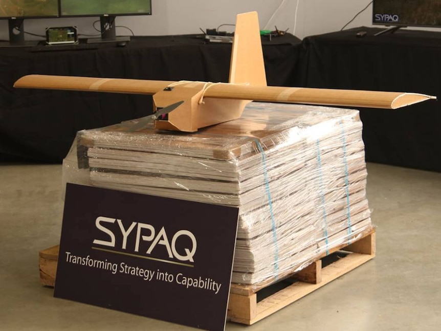 A cardboard drone sits atop a palette of flat-packed drones bearing a black sign that says 'SYPAQ'