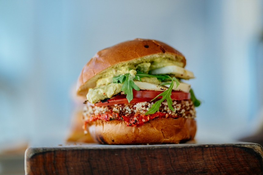  A burger in a bun with green avocado, green lettuce, red tomato and sauce and brown patty sits on a wooden chopping board