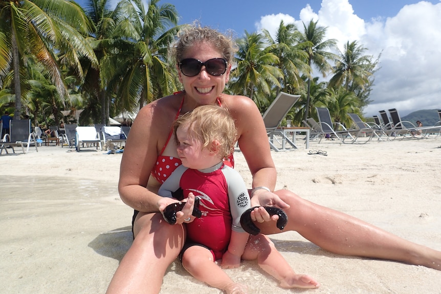 Woman holds black sea cucumbers on beach with young sitting between her legs.