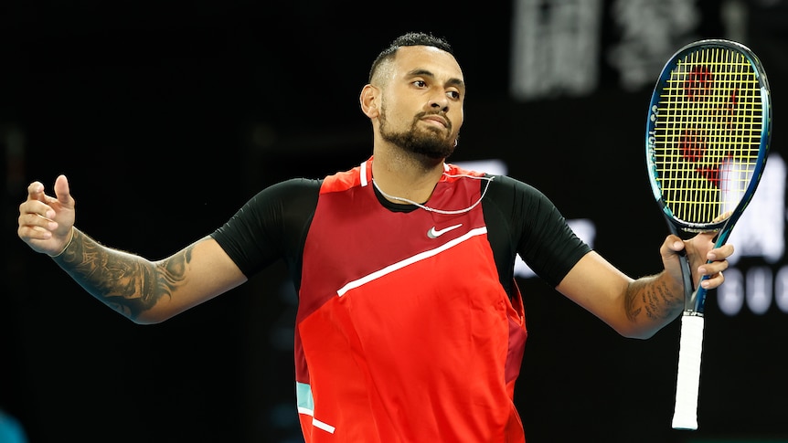 An Australian male tennis players stands holding his racquet with his arms outstretched.