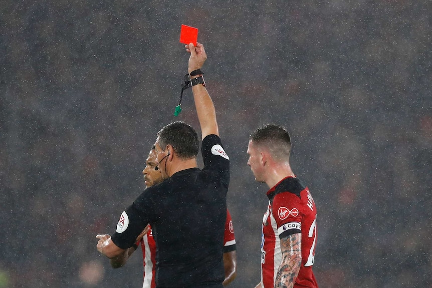 A Premier League referee holds up a red card in the rain to send off Southampton's Ryan Bertrand. Another player stands nearby.