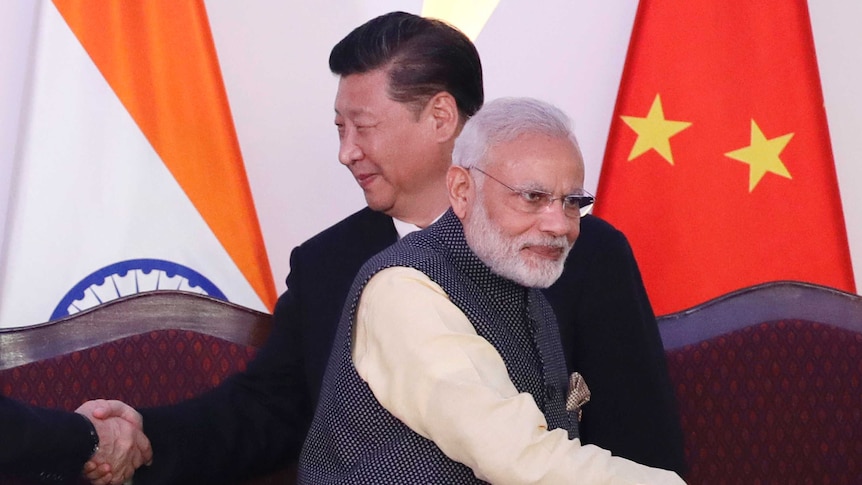 Two men stand and shake hands with two unseen people while standing in front of an Indian and a Chinese flag.