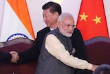 Two men stand and shake hands with two unseen people while standing in front of an Indian and a Chinese flag.