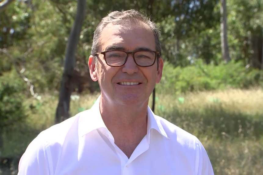 A man wearing black square glasses and a white button up shirt stands in front of grass and trees