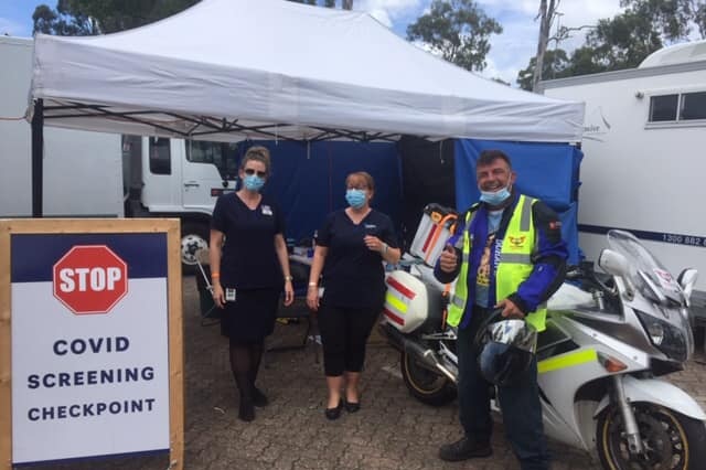 Two healthcare workers and a man smile for a photo at pop up testing clinic. The man is leaning against his motorcycle.