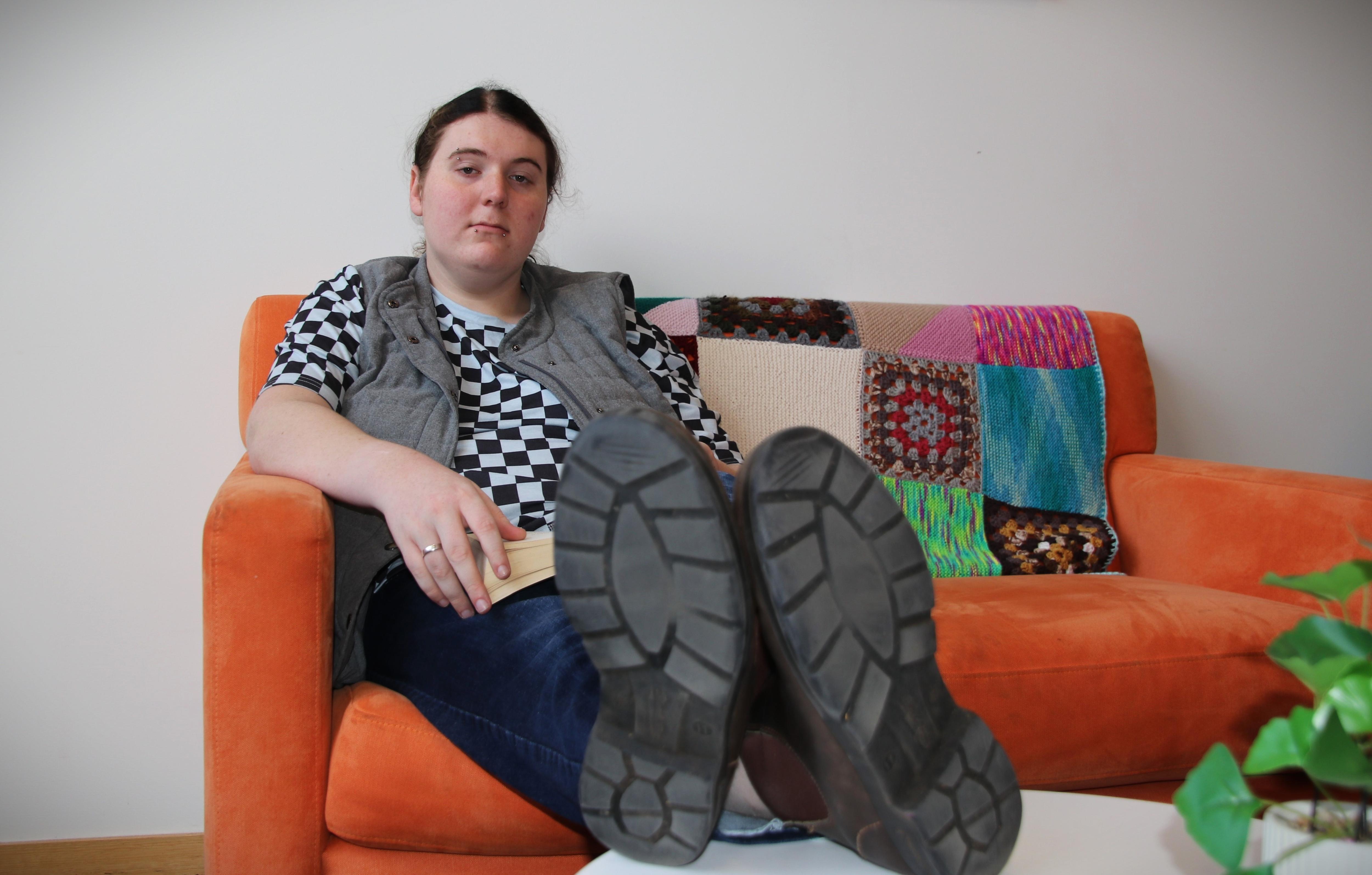 A former student stares down the barrel of the camera with a serious expression. She is sitting on an orange couch. 
