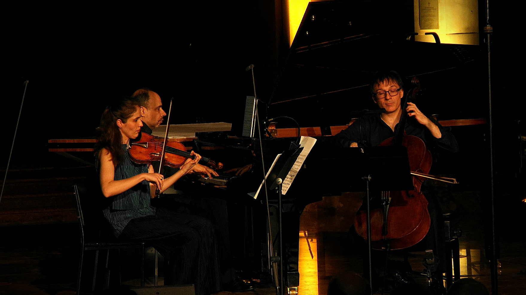 A female violinist with her hair tied back plays with a male pianist and a male cellist. Their expressions are concentrated.