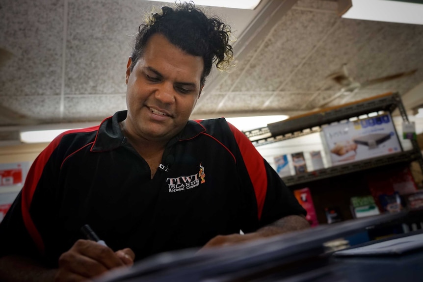 Jason de Santis smiles while writing on something behind a desk.