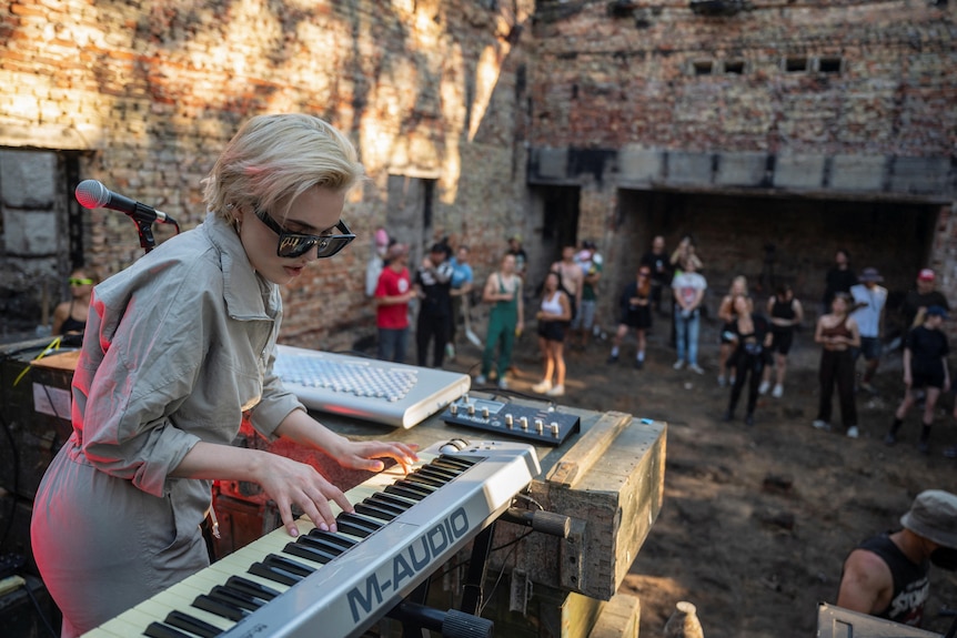 Женщина с короткими светлыми волосами, в солнечных очках играет на клавиатуре, пока люди смотрят в разрушенном здании