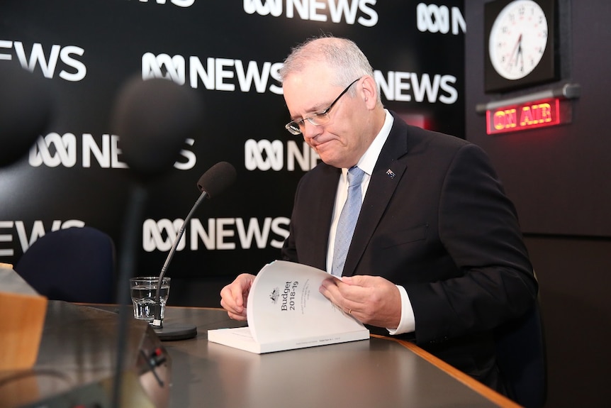 Scott Morrison looks at the Budget 2018-19 document while sitting in front of a microphone against an ABC News background.