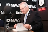 Scott Morrison looks at the Budget 2018-19 document while sitting in front o a microphone against an ABC News background