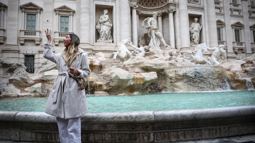 A woaman in a white coat with long brown hair prepares to toss a coin behind her into the Trevi Fountain