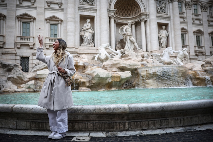 A woaman in a white coat with long brown hair prepares to toss a coin behind her into the Trevi Fountain