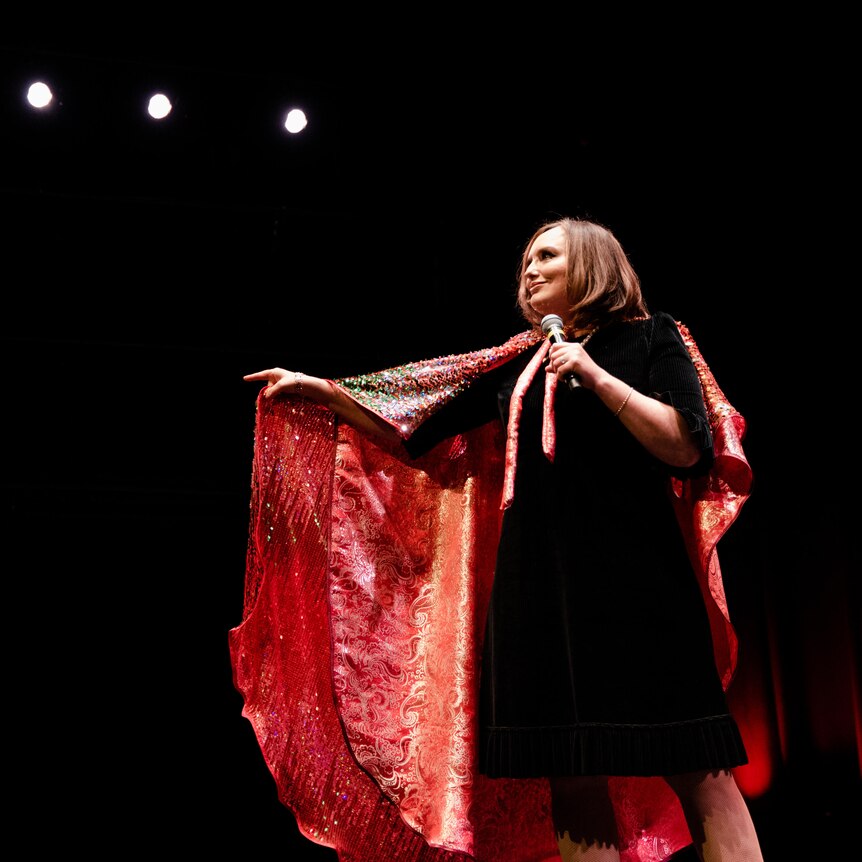 A woman stands on a dark stage, in one hand she holds a microphone, the other hand is pointing. She appears to be talking.