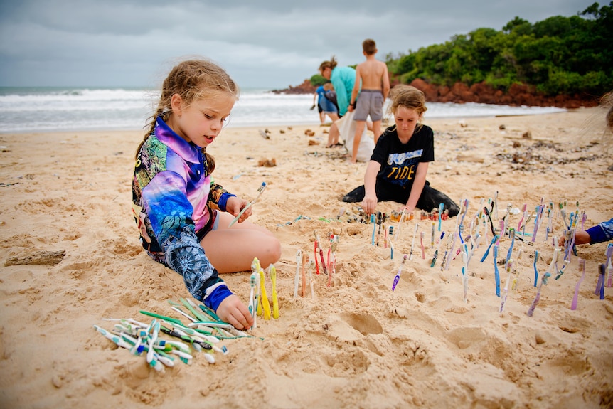 Two young children lining up toothbrushes that have washed up on a remote beach in the sand. 