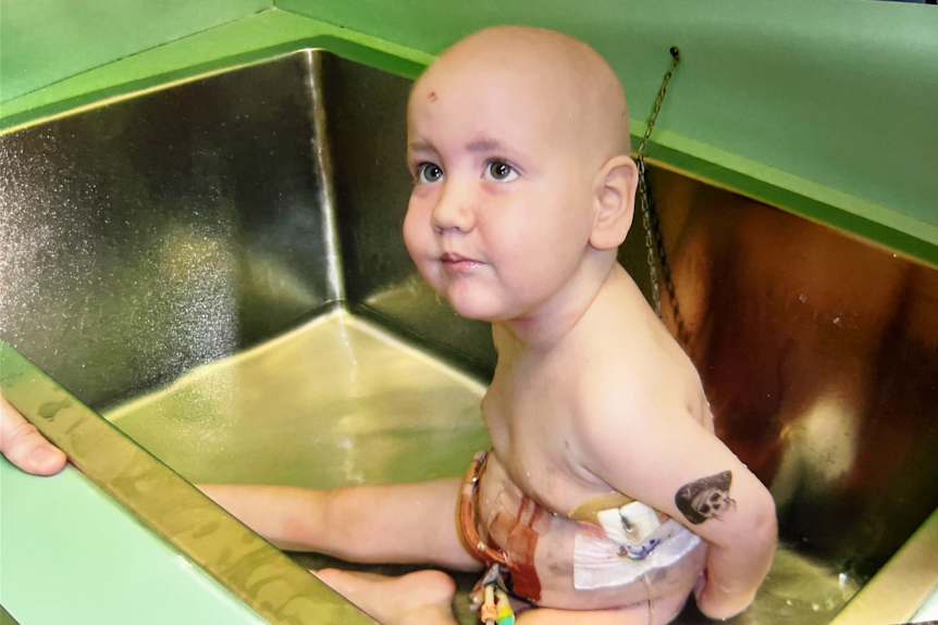Young boy sits in a metal sink with medical tubes attached to his body