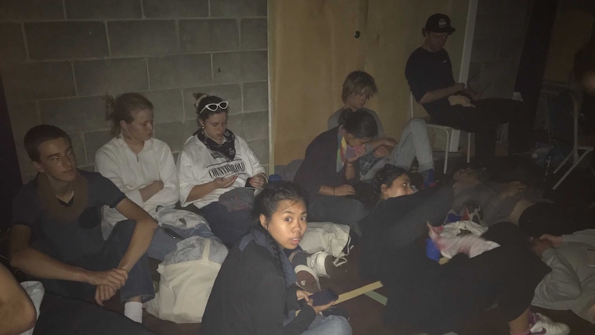 Ten young people sit and lie on the floor in a dark room.