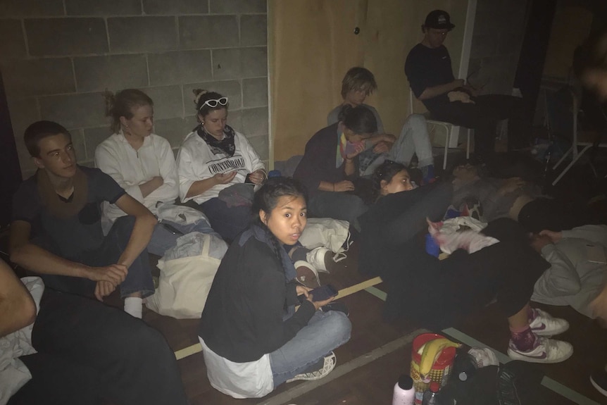 Ten young people sit and lie on the floor in a dark room.