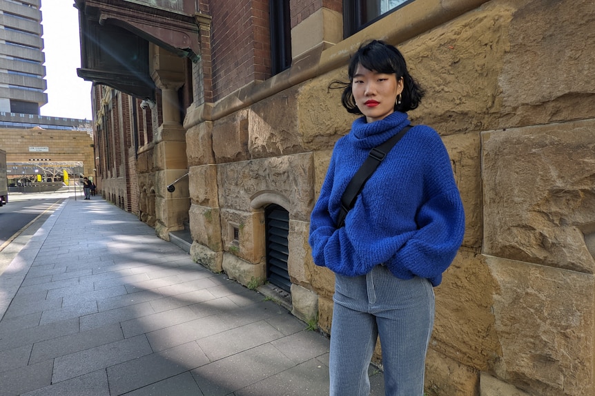 Chinese model with hands in pockets wearing blue jumper standing on street with sandstone in background.