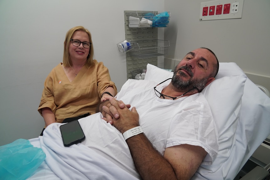 A man lies on a hospital bed, prepped to go into surgery, his wife by his side.