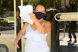 A woman wearing a white singlet top, floral skirt and black face mask holds up a piece of paper