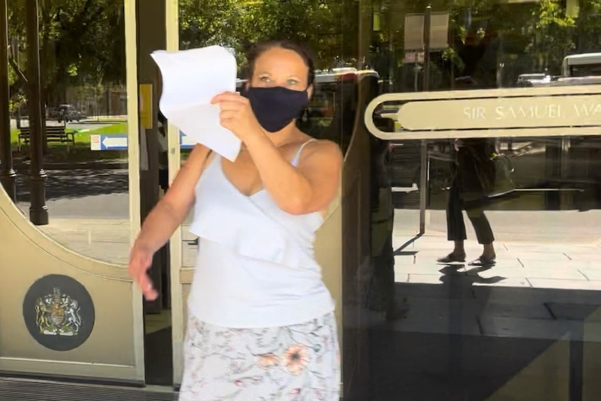 A woman wearing a white singlet top, floral skirt and black face mask holds up a piece of paper